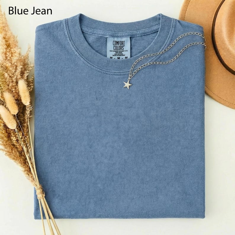 May include: A blue jean colored Comfort Colors t-shirt with a silver star pendant necklace and a brown hat. The t-shirt is folded and has a tag that reads "Comfort Colors". Dried wheat stalks are on the left side of the image.
