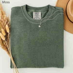 May include: A moss green t-shirt with a silver star pendant necklace. The shirt has a crew neck and a tag that reads "COMFORT COLORS". A tan hat and dried flowers are in the background.