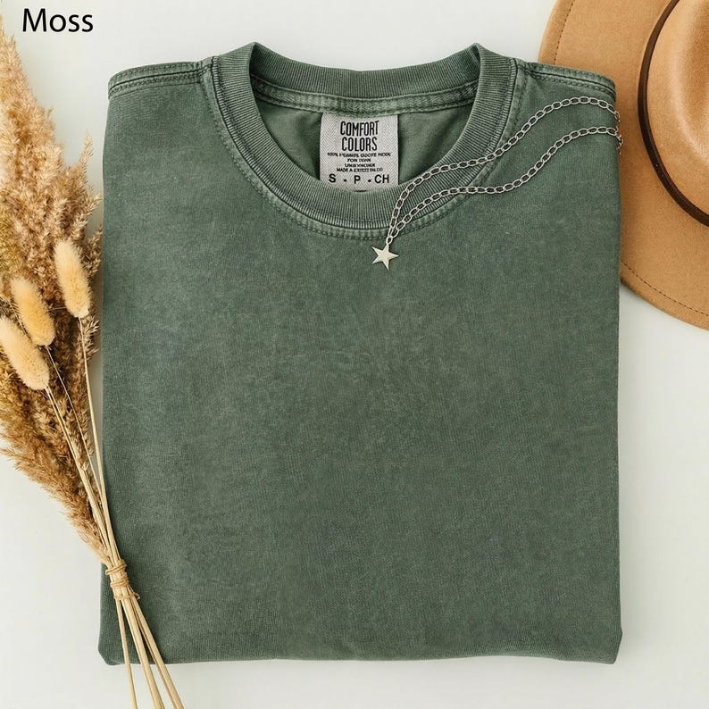 May include: A moss green t-shirt with a silver star pendant necklace. The shirt has a crew neck and a tag that reads "COMFORT COLORS". A tan hat and dried flowers are in the background.