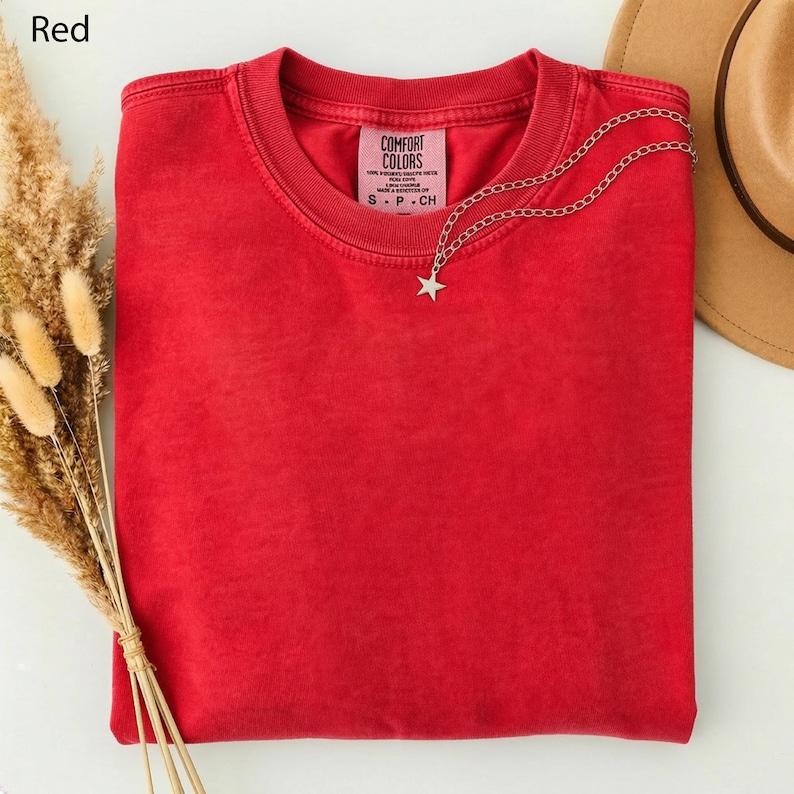 May include: A folded red Comfort Colors t-shirt with a silver chain necklace featuring a star pendant. The shirt has a crew neck and a small tag with text. The word "Red" is in the top left corner.