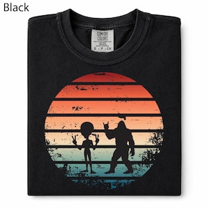 May include: Black crew neck t-shirt featuring a retro sunset design with an alien and a Bigfoot silhouette. The graphic includes peace signs and a rock-on hand gesture. The shirt has a Comfort Colors label.