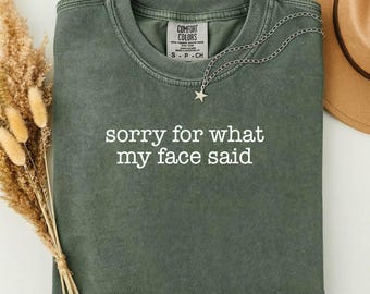 May include: A moss green t-shirt with the text "sorry for what my face said" in white. The shirt has a crew neck and a small tag at the neckline. A silver necklace with a star pendant is draped across the shirt.