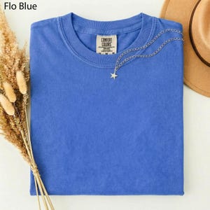 May include: A folded, blue Comfort Colors t-shirt with a silver star pendant necklace. The shirt has a crew neck and a tag that reads "Comfort Colors". The words "Flo Blue" are in the top left corner. A tan hat is in the background.