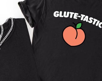 May include: Black t-shirt with the text "GLUTE-TASTIC" above a peach graphic. A silver chain necklace rests on a second black t-shirt. The shirts are made of a soft material.