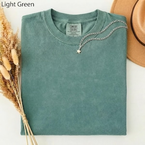 May include: A light green Comfort Colors t-shirt with a silver chain necklace featuring a star pendant. The shirt is folded and has a tag on the collar. The words "Light Green" are in the top left corner.