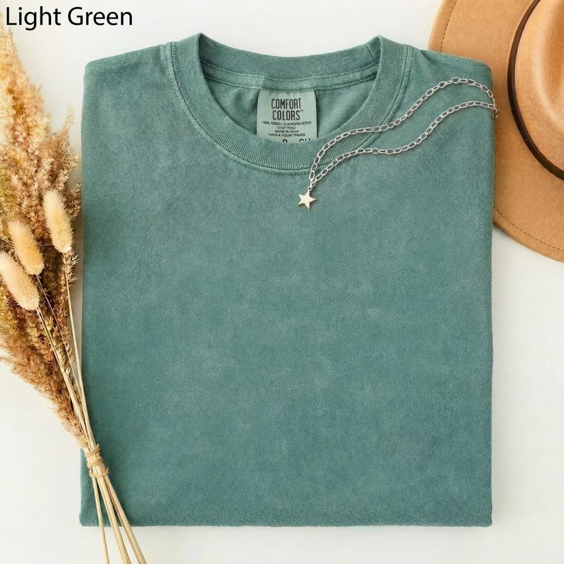 May include: A light green Comfort Colors t-shirt with a silver chain necklace featuring a star pendant. The shirt is folded and has a tag on the collar. The words "Light Green" are in the top left corner.
