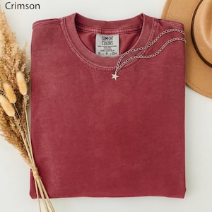 May include: Crimson-colored t-shirt with a crew neck, folded neatly. A silver necklace with a star pendant rests on the shirt. The tag reads "COMFORT COLORS". A tan hat is partially visible in the background.