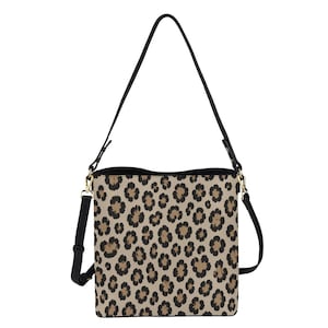 May include: A bucket-style shoulder bag featuring a tan base with a repeating black and dark brown leopard print pattern. The bag includes a black adjustable shoulder strap attached with gold-toned metal hardware clips on both sides.
