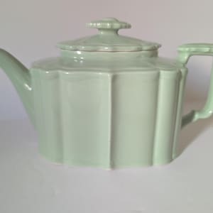 May include: A pale mint green ceramic teapot featuring a scalloped, fluted body design with vertical ridges. The teapot includes a matching lid with a small, decorative knob handle, a curved spout, and a rectangular handle on the side.