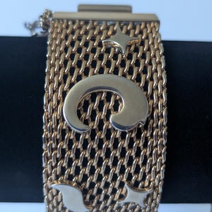 May include: A gold-toned mesh bracelet featuring a wide, flexible band with a woven texture. The surface is decorated with polished silver-toned metal accents, including a large central crescent shape, two four-pointed stars, and a curved element.