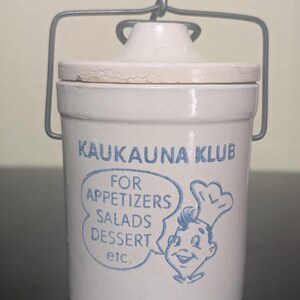 May include: A cylindrical off-white ceramic crock with a matching lid and a gray metal wire handle. The front features blue text reading KAUKAUNA KLUB, FOR APPETIZERS SALADS DESSERT etc., and a graphic of a person wearing a chef hat.