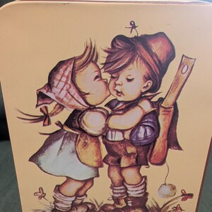 May include: A rectangular metal tin featuring a vintage-style illustration of a young girl in a checkered headscarf kissing a young boy wearing a hat and carrying a backpack. The artwork uses soft, muted tones of brown, cream, and blue.