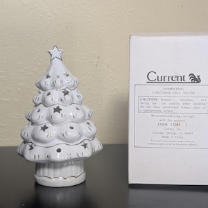 May include: A white ceramic Christmas tree votive candle holder with silver-trimmed star cutouts and a star topper, standing next to its original white box labeled Current Shimmering Christmas Tree Votive with caution and product information.