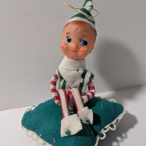 May include: A small elf figurine with a painted face, blue eyes, and rosy cheeks, wearing a green and white striped hat and matching outfit. The elf has red and white striped limbs and sits on a square, dark green felt cushion with white lace trim.