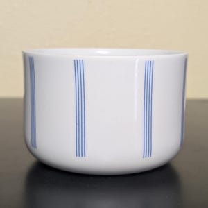 May include: A white ceramic bowl with a smooth, rounded silhouette. The exterior surface is decorated with vertical patterns consisting of four thin, parallel blue lines spaced evenly around the circumference of the bowl, creating a clean, simple design.