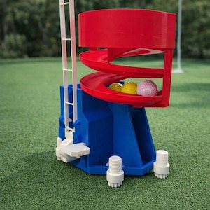 Puede incluir: Un sistema de retorno de bolas de golf azul y rojo con una pista en espiral. El sistema tiene un asa blanca y contiene tres bolas de golf, incluyendo amarillas y rosas. El dispositivo está sobre una superficie verde.