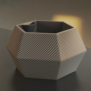 May include: A hexagonal planter with a light beige exterior featuring a series of diagonal, raised ridges across its six sides. The interior of the container is a dark charcoal gray, and the top rim has a smooth, flat edge with a small indentation.