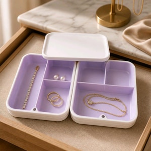 May include: A white and lavender jewelry organizer with a lid. The organizer is divided into compartments, holding a gold bracelet, rings, earrings, and a necklace. The jewelry box is on a beige surface.
