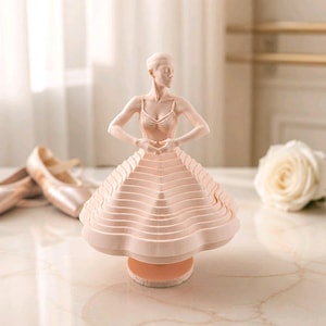 May include: A light pink ballerina figurine with arms gracefully posed. The dancer's layered skirt and base are the same colour. Ballet shoes and a white rose are in the background. The figurine is a decorative item.