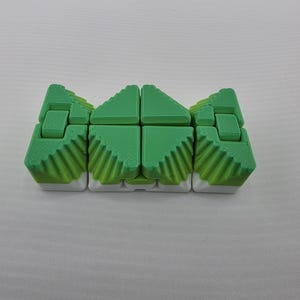 May include: A green and white fidget toy with a unique design. The toy is composed of several connected blocks, each with a textured surface and a distinct shape. The blocks are primarily green with white accents, creating a visually appealing contrast. The toy is designed to be manipulated.
