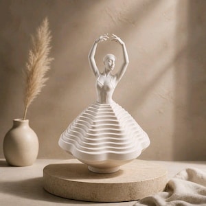 Op de afbeelding: Een witte keramische ballerinafiguur met opgeheven armen. De jurk van de danseres heeft gelaagde, golvende lagen. De sculptuur staat op een ronde, lichtgekleurde sokkel. Een decoratief object.