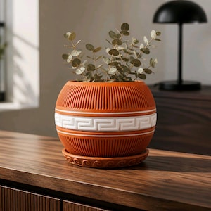 May include: A terracotta-coloured ceramic plant pot with a white Greek key pattern and matching saucer. The round pot has vertical grooves and contains a small plant. The pot sits on a wooden surface.