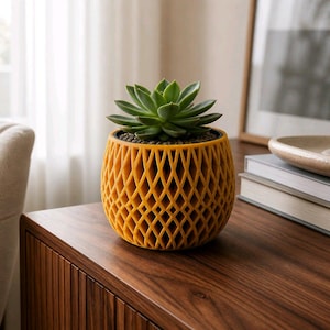 May include: A vibrant mustard yellow planter with a complex, repeating diamond-shaped lattice pattern. The round, spherical pot contains a green succulent plant with thick, pointed leaves arranged in a tight, symmetrical rosette formation.