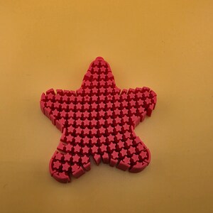 May include: A bright pink, five-pointed star-shaped object featuring a textured surface composed of numerous small, raised star-shaped protrusions covering the entire top face of the item, creating a uniform and repeating pattern across the surface.