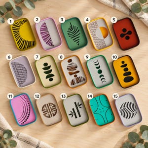 Puede incluir: Quince bandejas rectangulares dispuestas en tres filas, cada una con un diseño gráfico único. Las bandejas muestran varios colores, incluyendo amarillo, morado, verde, naranja y turquesa, con patrones negros o blancos como soles y lunas.