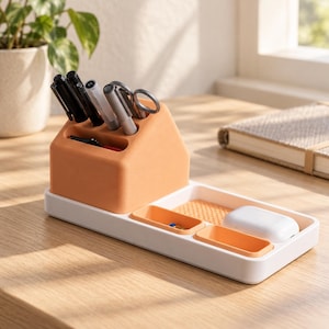 May include: A peach-colored desk organizer with a house-shaped pen holder section and a white rectangular base. The base features two small peach-colored compartments and a textured tray area holding a white wireless earbud charging case.