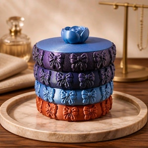 Op de afbeelding: Een cilindrische, stapelbare opbergdoos met vier afzonderlijke secties in tinten paars, blauw en oranje. Elke sectie is voorzien van een herhalend reliëfpatroon van strikjes aan de buitenkant. Een blauwe, roosvormige knop zit op het deksel.