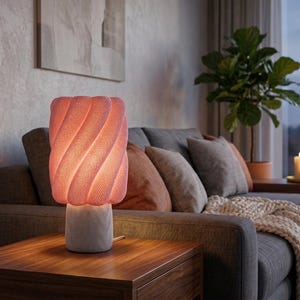 May include: A table lamp featuring a cylindrical white base with a textured surface and a large, pink, spiraling, ribbed lampshade that emits a soft, warm glow. The lamp is positioned on a wooden surface, showcasing a distinct, modern, and sculptural design.