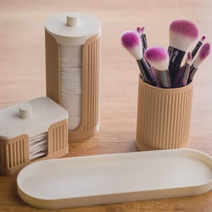 May include: A beige and white bathroom vanity set featuring a cylindrical cotton pad dispenser, a rectangular cotton swab holder, a cylindrical makeup brush cup filled with brushes with pink-tipped bristles, and an empty oval tray.