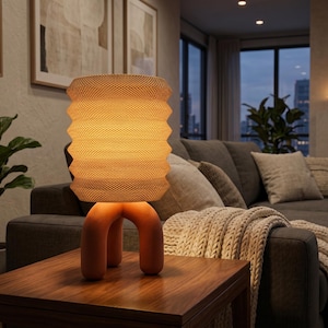 Peut inclure: Une lampe de table dotée d'un abat-jour cylindrique texturé à plis accordéon dans une teinte jaune chaud, reposant sur une base lisse de couleur terre cuite avec trois pieds arrondis en forme d'arche assurant la stabilité du luminaire.