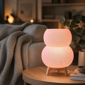 May include: A table lamp with a double-bulb, stacked spherical shape featuring a fine, white, grid-like textured pattern. The lamp emits a warm, soft glow and is supported by three light-colored wooden legs, creating a stable base on a flat surface.