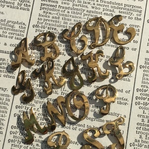 Puede incluir: Un conjunto de abalorios de letras del alfabeto de metal de color dorado. Las letras están en estilo de escritura cursiva. Las letras están sobre un fondo blanco con texto negro.