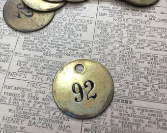 May include: A brass tag with the number 92 printed in black. The tag has a hole at the top for hanging. The tag is surrounded by other brass tags with different numbers.