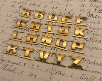 May include: Gold-colored metal alphabet letter stamps with a raised design. The letters are arranged in alphabetical order on a piece of paper with handwritten numbers and text.
