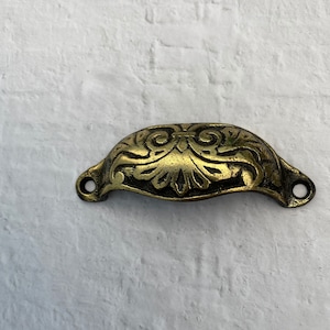 May include: A gold-toned metal cabinet pull with an ornate, floral design. The pull is shaped like a half-circle and has two small holes on either side for attaching to a cabinet door.