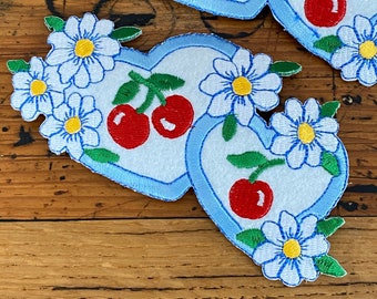 May include: Two blue felt hearts with embroidered white daisies and red cherries. The hearts are stitched together and have a light blue outline.