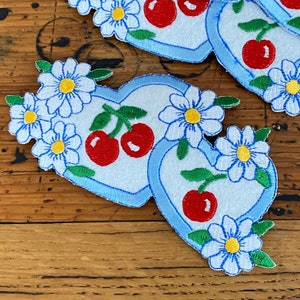 May include: Two blue felt hearts with embroidered white daisies and red cherries. The hearts are stitched together and have a light blue outline.