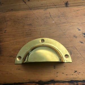 May include: A shiny, half-moon-shaped brass drawer pull with a curved handle. The hardware has three pre-drilled holes for mounting. The pull is a bright gold color and reflects light.