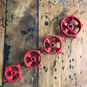 Puede incluir: Cuatro volantes de válvula rojos, con centro cuadrado y radios redondeados, dispuestos diagonalmente sobre una superficie de madera envejecida. Los volantes muestran signos de desgaste, con pintura saltada, lo que añade encanto antiguo.
