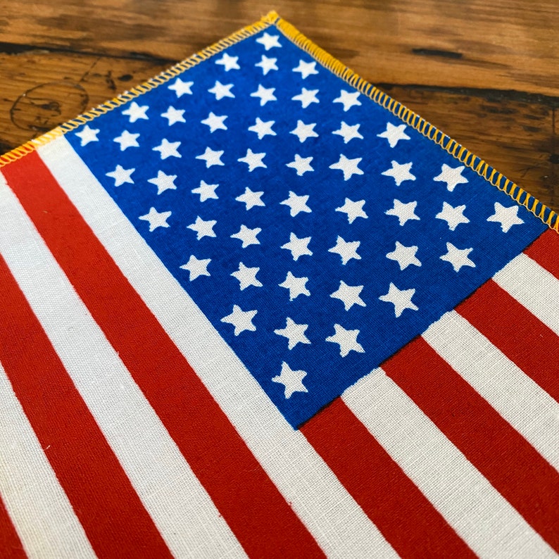 Pu&ograve; includere: Un primo piano di una piccola bandiera americana con strisce rosse e bianche e un campo blu di stelle. La bandiera &egrave; fatta di tessuto e ha un bordo cucito giallo.