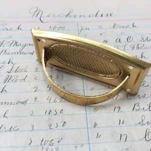 Könnte beinhalten: Ein goldfarbener Metall-Schubladenzug mit einer ovalen Öffnung und einer dekorativen, strukturierten Oberfläche. Der Zug hat einen gebogenen Metallgriff.