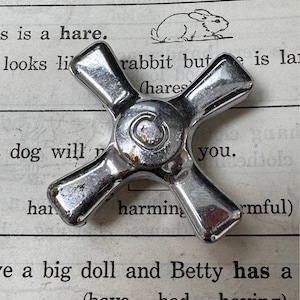 May include: A silver cross-shaped faucet handle with a round knob in the center. The handle is on a page of a book with text about a hare.