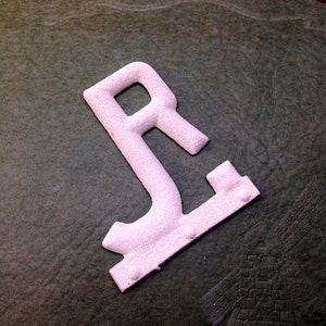 Puede incluir: Una letra "R" blanca en relieve sobre una superficie verde oscuro. La letra está hecha de un material texturizado y tiene un aspecto ligeramente desgastado.