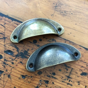 May include: Two antique-style cabinet pulls, one in a brass finish and the other in a dark bronze finish. Both pulls have a rounded, cup-like shape.