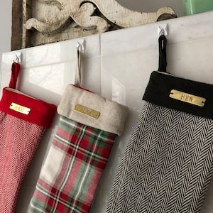 Puede incluir: Tres calcetines de Navidad con etiquetas de nombre de latón personalizadas. Los calcetines son de cuadros rojos y blancos, de espiga negra y blanca y de espiga roja y blanca. Los nombres en las etiquetas son Alex, Ben y Jenni.