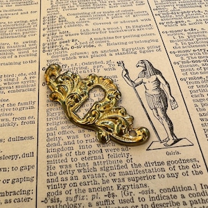 May include: A gold-coloured metal decorative accent with intricate floral designs. The accent is shaped like a stylised leaf with a hole in the centre. The accent is on a page of a book with text about the Egyptian god Osiris.